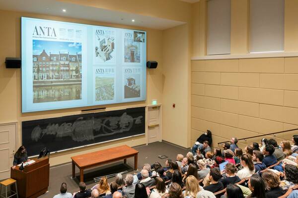 A woman presents at a lectern to an audience in a lecture hall.  Behind her, a partially erased chalkboard and a projected image of the Archives of New Traditional Architecture journal cover multiple pages.