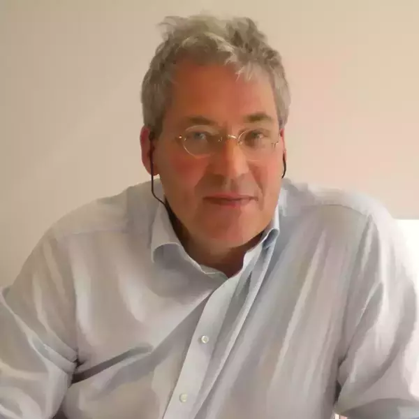 A man with gray hair and gold-rimmed glasses wears a light blue shirt. He has a black earpiece in his right ear and a calm expression, looking forward.