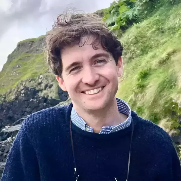 A man with wavy brown hair and blue eyes smiles broadly, wearing a dark blue sweater over a striped shirt. He stands against a lush green, rocky hillside under a cloudy sky.