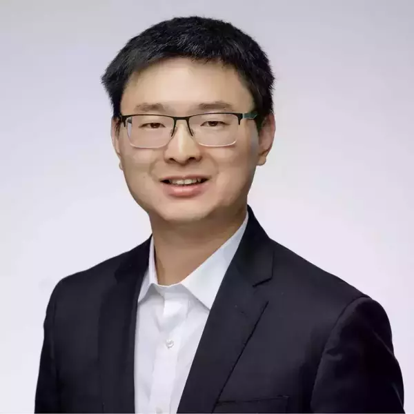 A man with short black hair and dark-rimmed glasses smiles slightly while wearing a black suit jacket and white shirt.