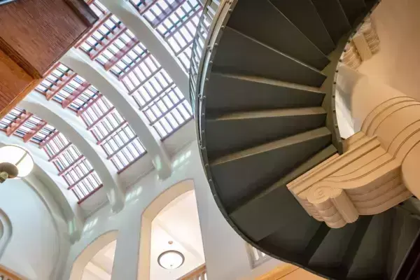 Interior view looking up inside the Royal College of Music. A dark gray spiral staircase curves upwards, supported by an ornate light beige column. Overhead, arched skylights with dark wood frames illuminate the grand interior with light-colored walls and arches.