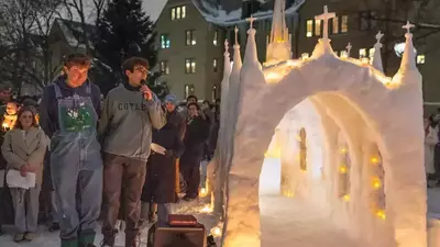 At night on Notre Dame's snowy campus, students gather around an elaborate snow church sculpture with arched entrances, spires, and crosses. Many small candles glow within. Two young men, one speaking into a microphone, stand prominently.