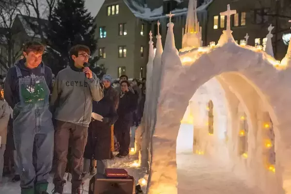 At night on Notre Dame's snowy campus, students gather around an elaborate snow church sculpture with arched entrances, spires, and crosses. Many small candles glow within. Two young men, one speaking into a microphone, stand prominently.