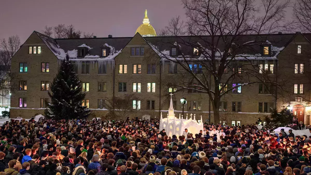 A large crowd gathers, holding lit candles at night in front of Zahm Hall, which is covered in snow and icicles. A white snow sculpture of a church stands among them. The Golden Dome shines in the background.