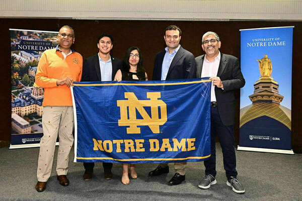 Five smiling people, including a woman and four men, hold a blue Notre Dame flag with gold ND logo and "NOTRE DAME" text.