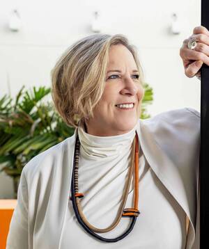 Smiling woman (Joyce Owens), blonde hair, blue eyes, in cream clothing, with black, brown, orange necklace. Hand with silver ring on dark column.