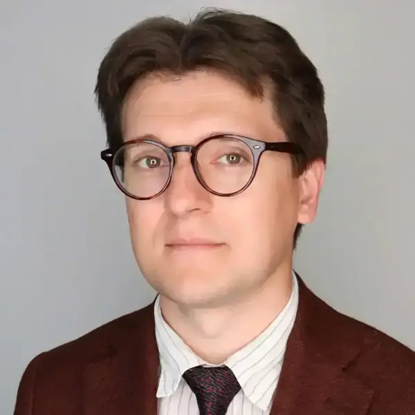 Man with brown hair and tortoiseshell glasses, wearing a russet jacket, striped shirt, and patterned tie. Neutral expression.