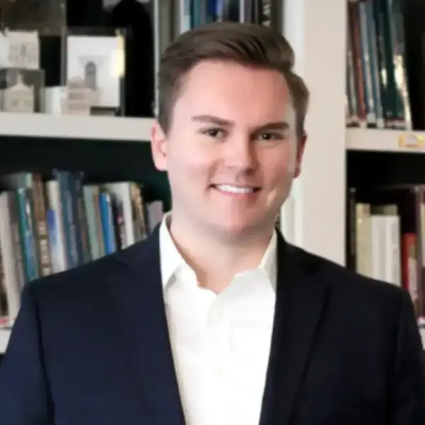 A smiling man with light brown hair in a navy blazer and white shirt against a blurred bookshelf.