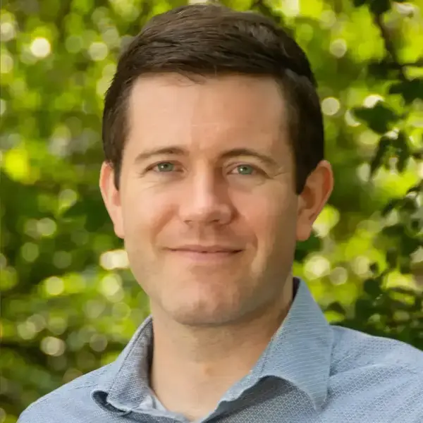 A smiling man with brown hair and blue eyes wears a light blue collared shirt against a blurry green background.