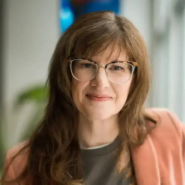 A woman with brown hair and tortoise-shell glasses smiles gently, wearing a peach jacket over a dark shirt.