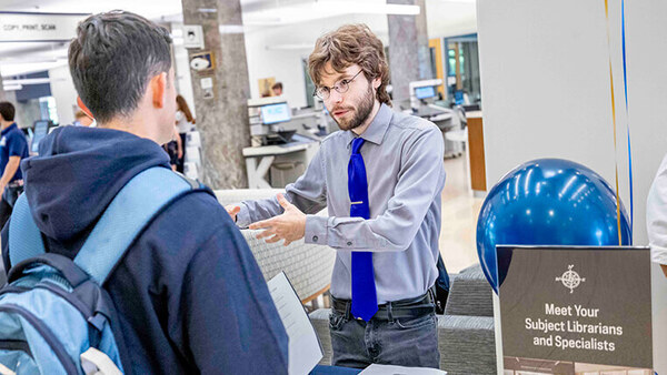A faculty member standing behind a table and facing the camera gestures while speaking with a student wearing a backpack. A sign on the table says "Meet Your Subject Librarians."