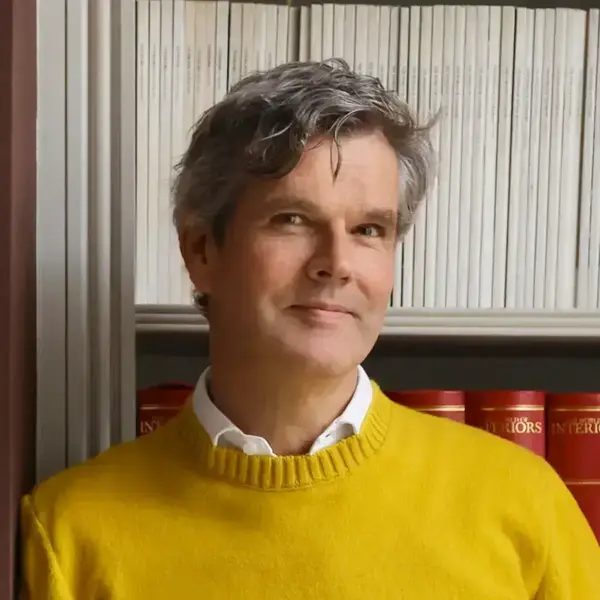 A man with gray hair, wearing a yellow sweater, smiles gently in front of a bookshelf stocked with white and red books.