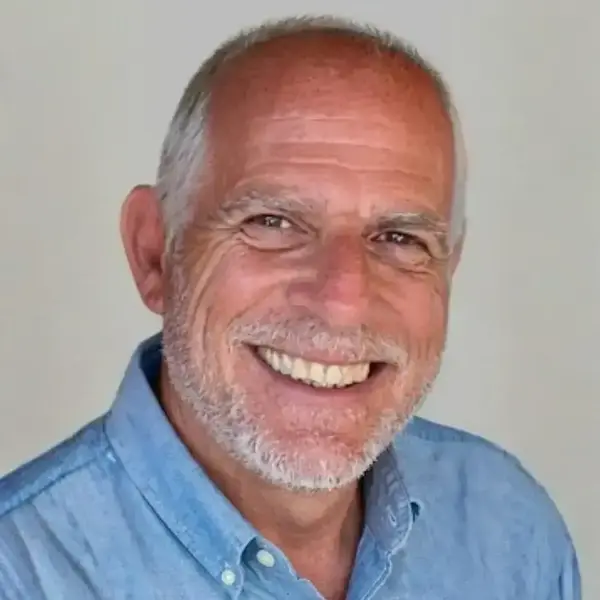 A man with gray hair and beard, wearing a light blue shirt, smiles widely, showing teeth against a plain background.