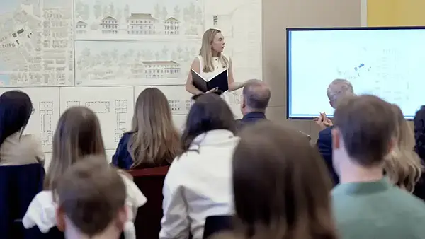 A blonde woman in white presents architectural plans to an audience in a classroom with drawings on the wall and screen for her fifth-year thesis project.