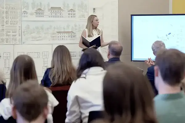 A blonde woman in white presents architectural plans to an audience in a classroom with drawings on the wall and screen for her fifth-year thesis project.