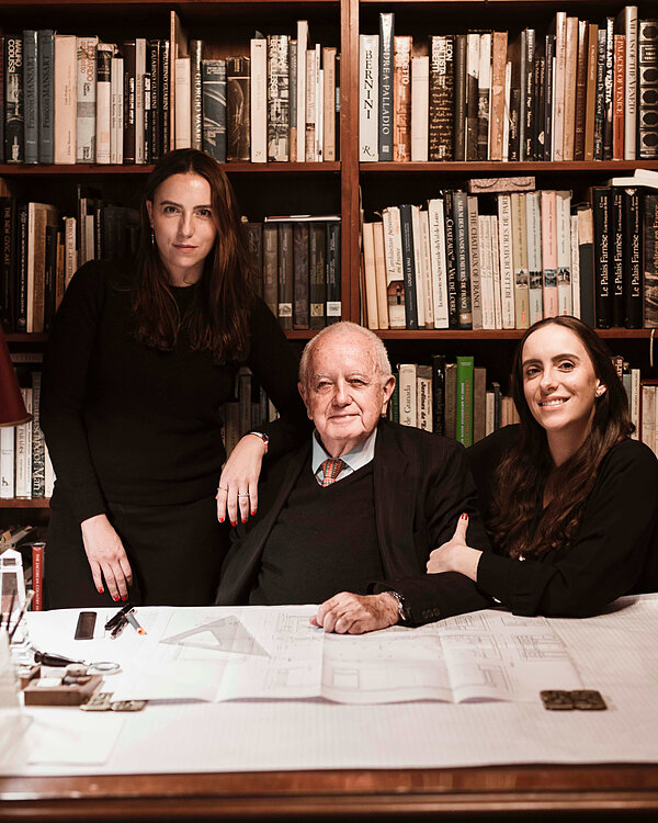 Older man at blueprint desk, flanked by two women. One stands serious behind him, another leans smiling. Books of architecture behind.