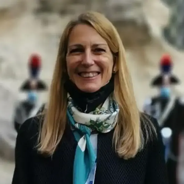 Smiling blonde woman wears a black top and blue-green patterned scarf. Blurred background has two guards with red plumed helmets.