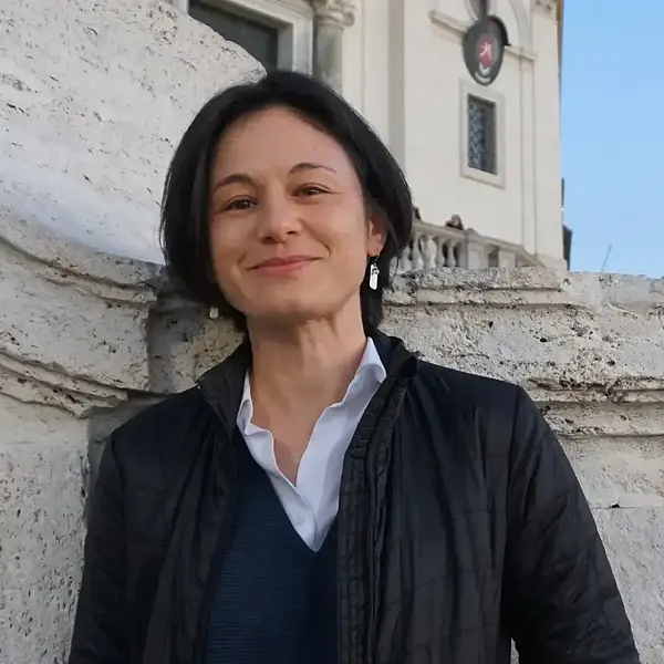 Woman with short dark hair smiles, wearing a black jacket over a blue sweater and white shirt.